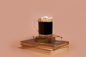 A glass cup of coffee with marshmallows and glasses on a stack of old books