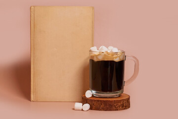 A glass cup of coffee with marshmallows stands on a wooden stand next to book