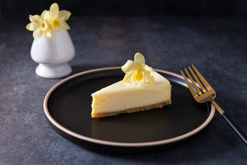 Classic New York cheesecake with fresh vanilla flower on dark concrete background, side view. A piece of Vanilla cheesecake on a black plate. Confectionery menu, recipe. Close up