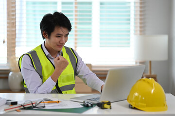 Young asian engineer worker in reflective jacket looking at laptop computer and celebrating success.