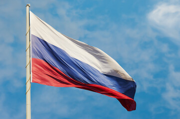 The national flag of the Russian Federation flutters in the wind against a blue sky with white clouds. The tricolor symbol of the state, attached to a pole with a cord.