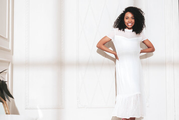 Beautiful black woman with afro curls hairstyle. Smiling model dressed in white summer dress. Sexy carefree female posing near wall in studio. Tanned and cheerful. At sunny day. Shadow from window