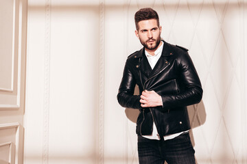 Handsome confident  model. Sexy stylish man in biker leather jacket and black jeans. Fashion hipster male posing near white wall in studio interior at sunny day. Shadow from window