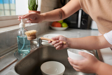 Hands of mother helping son to add dishwashing detergent on brush when he is cleaning bowls after breakfast