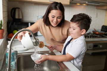 Smiling mother helping son to rinse dishes under tap water after cleaning