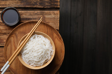 Bowl with cooked rice noodles, soy sauce and chopsticks on wooden table, flat lay. Space for text