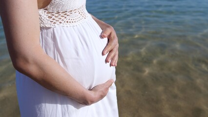 Fototapeta na wymiar A pregnant woman in a white dress on the seashore stands and strokes her tummy, communicates with the baby in her stomach. Walking in the fresh air for a good pregnancy.