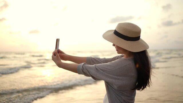 Young asian woman using smartphone to take a photo of seaview while sunset time. Traveler female Relaxing on holiday weekend vacation time. Pick up cell phone to capture the impression