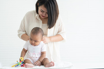 Adorable African newborn girl sitting and holding toy in hand while young Asian mother dressing changing her infant clothes after bath at home. White background. Bonding in mixed race family
