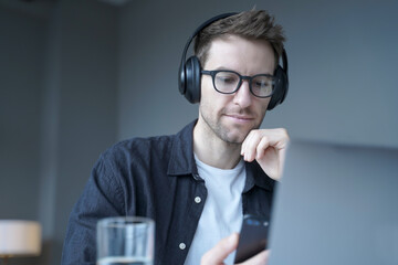 Young focused german man in wireless headphones using mobile phone listening music at workplace
