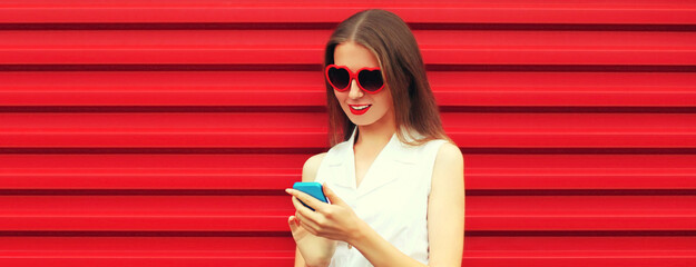 Portrait of young woman with phone wearing red heart shaped sunglasses on background
