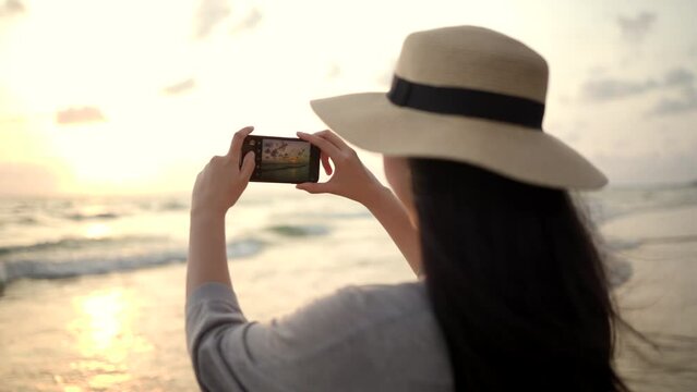 Young asian woman using smartphone to take a photo of seaview while sunset time. Traveler female Relaxing on holiday weekend vacation time. Pick up cell phone to capture the impression