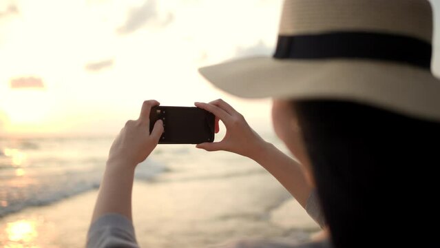 Young asian woman using smartphone to take a photo of seaview while sunset time. Traveler female Relaxing on holiday weekend vacation time. Pick up cell phone to capture the impression