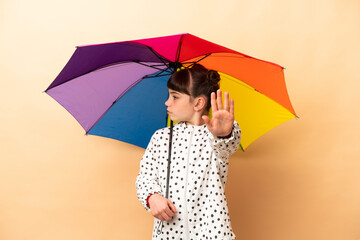 Little girl holding an umbrella isolated on beige background making stop gesture and disappointed