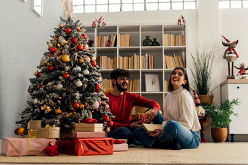 Happy woman with boyfriend holding gift box sitting by Christmas tree at home