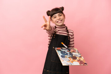 Little artist girl holding a palette isolated on pink background smiling and showing victory sign