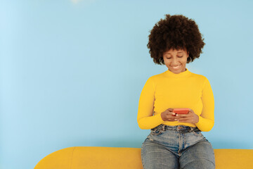 Smiling woman surfing net through mobile phone sitting on yellow sofa against blue background