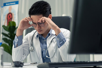 Stressed and overworked Asian male doctor sitting on table in hospital. 