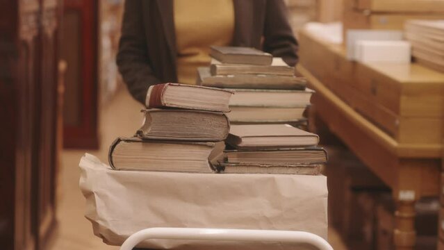 Midsection slowmo of unrecognizable female librarian carrying old rare books in cart while walking along drawers with card catalog in library