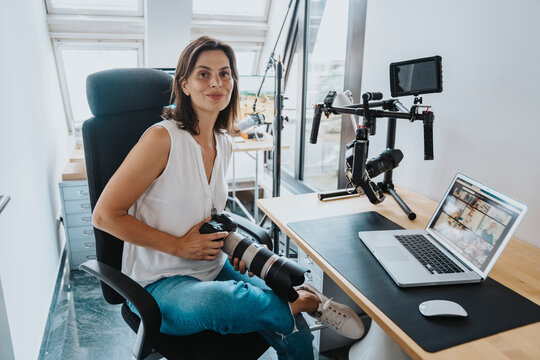 Smiling photographer with digital camera sitting on chair with laptop on table at studio