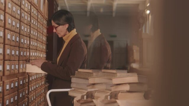 Medium slowmo of mature Caucasian female librarian in eyeglasses standing by drawers with library paper database or archive searching for necessary book