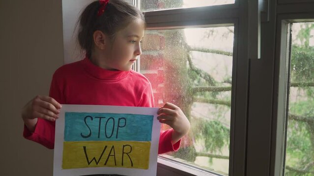 Refugee girl looking outside window and holding banner with inscription Stop war at blue yellow flag of Ukraine . Crisis, peace, stop aggression in whole world