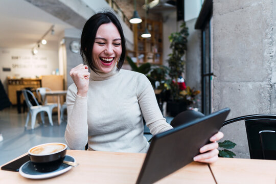 Cheerful woman gesturing fist holding tablet PC sitting in cafe