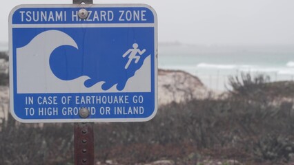 Ocean beach sandy dunes, Monterey, California misty coast, USA. Foggy rainy autumn, winter weather, grey cloudy sky. Trail path on shore, cold sea waves. Warning sign, tsunami hazard. Danger caution.