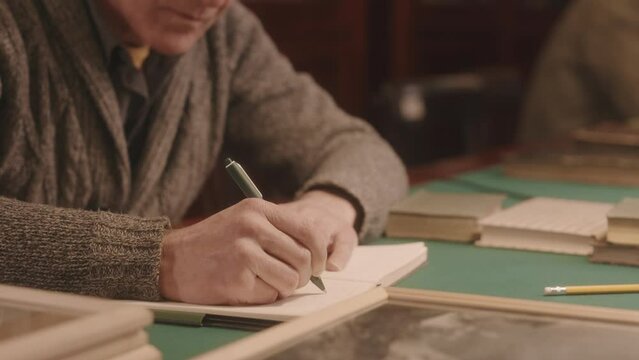 Cropped slowmo of Caucasian senior man sitting at wooden desk in library taking notes in notebook