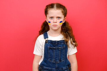 Portrait of little girl with flag of ukraine on face, looking at camera, isolated on red studio background. Peace, no war, help, stop russian aggression. Evacuation of civilians. Freedom to Ukraine