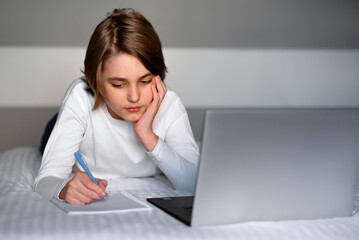 Teenager boy writing at notebook in front of laptop, sitting on bed. Boy having lessons, studying online at home. Distance education concept. Home interior.