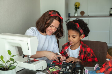 Caucasian mom and an African-American daughter sew clothes together for a doll at home on a sewing machine.Diverse people.Time together.Mother's day.Selective focus. - obrazy, fototapety, plakaty