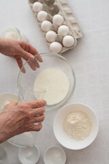 Process of making dough cake. woman’s hands pours milk in bowl. On the side stand eggs, flour and  sugar on white table. ingredients for baking cake in kitchen, recipe. Top View.