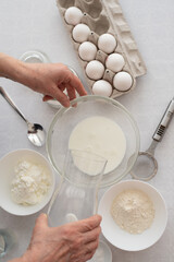 Process of making dough cake. woman’s hands pours milk in bowl. On the side stand eggs, flour and  sugar on white table. ingredients for baking cake in kitchen, recipe. Top View.