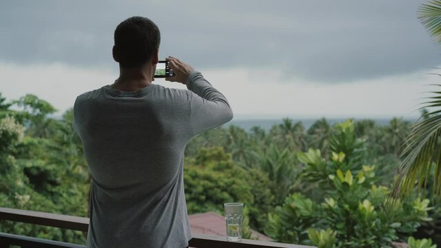 A man stands on a balcony and taking pictures on smartphone. Tropical greenery, sea and rain clouds in the background.