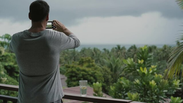 A man stands on a balcony and taking pictures on smartphone. Tropical greenery, sea and rain clouds in the background.