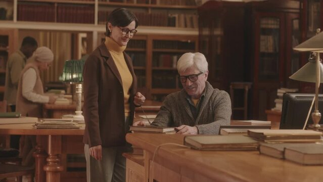 Medium slowmo of mature Caucasian woman borrowing book from public library asking senior librarian sitting by laptop at wooden reception desk