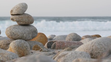 Rock balancing on pebble beach, Monterey 17-mile drive, California coast, USA. Stable pyramid stacks of round stones, sea ocean water waves crashing at sunset. Serenity harmony, calm zen meditation.