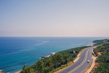 An empty asphalt road winds along the breathtaking scenic coastline on a sunny summer day. A spectacular shot of a coastal road overlooking clear blue skies and the calm Mediterranean Sea.