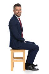 happy young man in elegant suit sitting in a side view pose on wooden chair