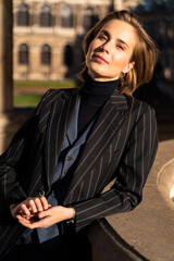 Young blond cheerful happy smiling business woman with pierced nose and earrings in retro styled striped jacket looking in camera leaned on historic stone building, in Dresden Zwinger on sunny day. 