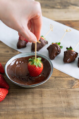 Strawberries with chocolate on a wooden background. Hand dipping a strawberry in the bowl of chocolate. Vertical photo.
