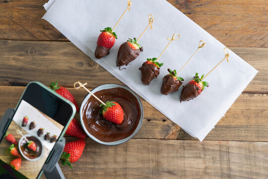 Strawberries with chocolate on a wooden background. Hand taking pictures with cell phone. Copy space.