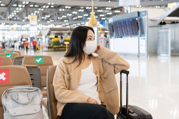 Beautiful Asian girl wearing medical mask sitting at international airport while waiting for check in counter open.  Female passenger at terminal, indoors. Health care, traveling concept