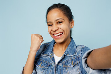 Close up cool little kid teen girl of African American ethnicity 12-13 years old in denim jacket do selfie shot pov on mobile phone do winner gesture isolated on pastel plain light blue background.