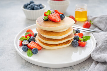 A stack of delicious fluffy pancakes with berries and honey on a white plate on a gray concrete background. Copy space.