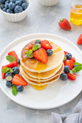 A stack of delicious fluffy pancakes with berries and honey on a white plate on a gray concrete background. Copy space.