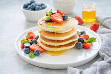 A stack of delicious fluffy pancakes with berries and honey on a white plate on a gray concrete background. Copy space.