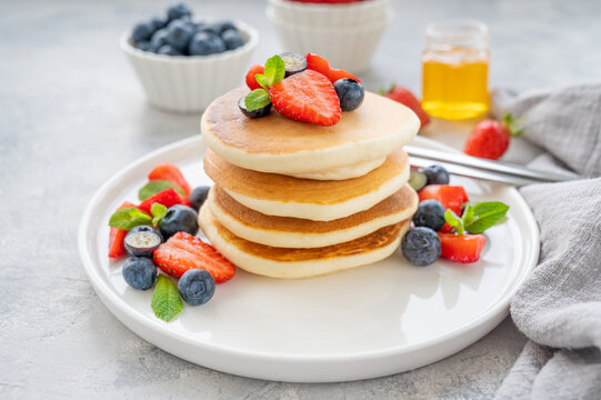 A Stack Of Delicious Fluffy Pancakes With Berries And Honey On A White Plate On A Gray Concrete Background. Copy Space.