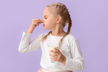 Cute little girl with glass of milk closing nose on lilac background